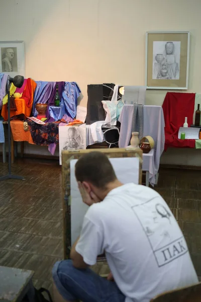 A student of an art school in a painting lesson at a tablet, plans a drawing of a staged still life of draperies, earthenware, glass bottles, a yellow teapot, in front of drawings in glass frames hung on the wall