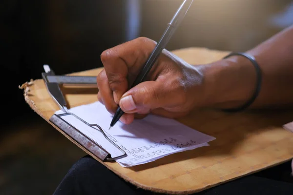 A man\'s hand is writing on a document with a board