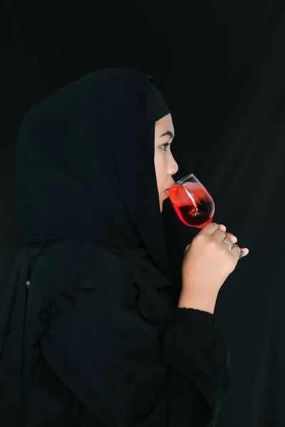 Muslim woman wearing a black dress drinking a glass of red syrup on black background