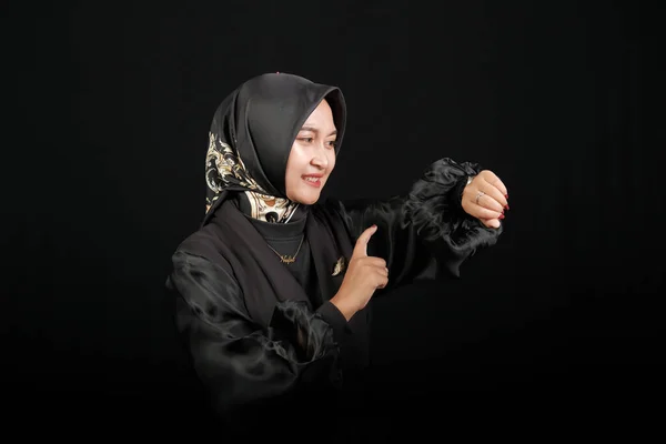 身穿黑色礼服的快乐的亚洲穆斯林妇女 戴着眼镜 看着手表 在黑屏的背景下微笑着 — 图库照片