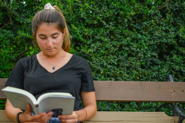 Genç beyaz bir kadının parkta bir bankada kitap okumasını ön planda görmek. Yatay olarak. Kopyalama alanı ile.