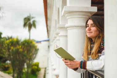 Beyaz bir kadın gülümsüyor ve elinde sütunlarla balkonda bir defter tutuyor. Yatay olarak. Kopyalama alanı ile.