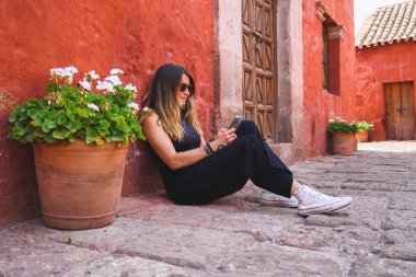 Orta çağ caddesinde kırmızı bir duvarda otururken gülümseyen ve cep telefonunu kontrol eden beyaz bir kadın. Yatay olarak. Kopyalama alanı ile.