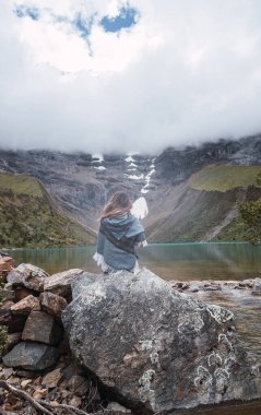 Beyaz bir kadının Humantay Gölü 'nün önündeki bir kayanın içinde tipik Peru pançosu giymiş bir şekilde oturması. Dikey. Kopyalama alanı ile.