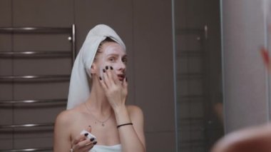 Kafasına havlu sarılmış bir kadın banyo aynasından önce kilden maske takıyor. Günlük akşam rutini - yüz temizleme, cilt bakımı, kabuk soyma, nemlendirici ve güzellik tedavi konsepti