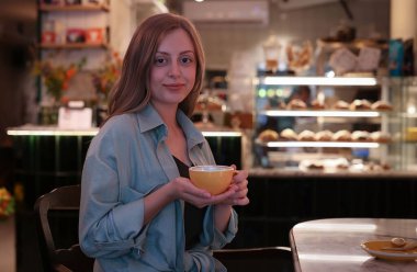 A girl sits in a cafe and holds a cup in her hands, looks at the camera