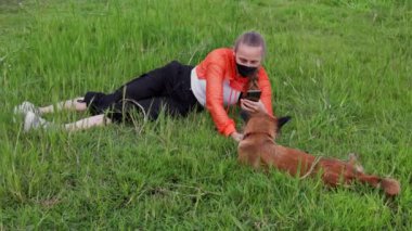 Saçlı, turuncu kapüşonlu, yüzünde maske olan sarışın bir kadın kırmızı bir köpekle çimenlerde oynuyor. Yüksek kalite 4k görüntü