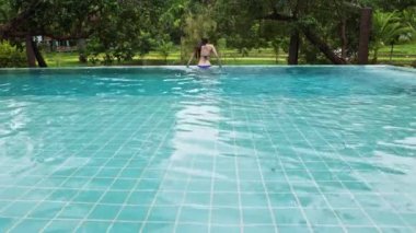 A woman sits on the edge of the pool and swims against the backdrop of tropical vegetation in a hotel or villa. High quality 4k footage