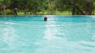 A beautiful long-haired woman swims and sits on the edge in an outdoor infinity pool against the backdrop of tropical vegetation. High quality 4k footage