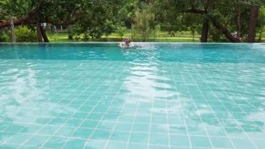 A woman sits on the edge of the pool and swims against the backdrop of tropical vegetation in a hotel or villa. High quality 4k footage