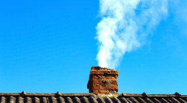 Parlak, doymuş bir gökyüzüne karşı ısınma sırasında bir kır evinin tepesindeki eski tuğla bacadan çıkan beyaz duman..