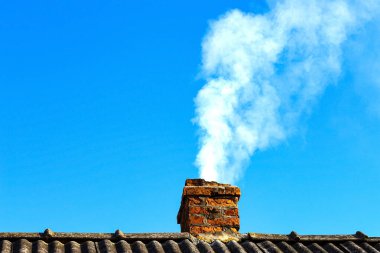 Parlak, doymuş bir gökyüzüne karşı ısınma sırasında bir kır evinin tepesindeki eski tuğla bacadan çıkan beyaz duman..