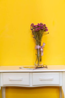 Çiçekli bir vazonun parlak kompozisyonu, sarı bir duvara karşı beyaz bir sehpanın üzerinde bir dergi ve bardaklar.