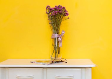 Kahve masasında çiçekli vazonun olduğu minimalist bir kompozisyon. Bir dergi ve gözlük var. Duvar rengi sarı. dikey fotoğrafçılık