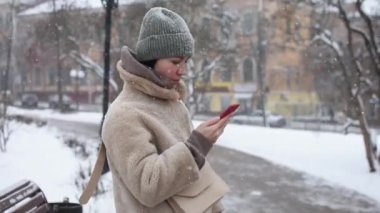 Kürk mantolu kız, kış parkının arka planındaki akıllı telefon kamerasıyla fotoğraf çekiyor. Kar yağıyor, soğuk hava