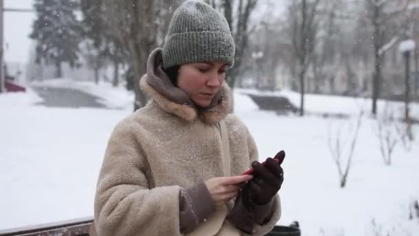 一个穿着毛皮外套的女孩正在一个冬季公园的背景下 用智能手机打字 下着雪 天气很冷 — 图库视频影像