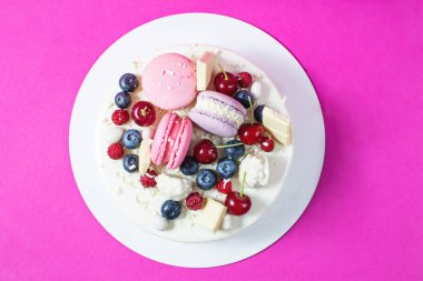 Tender white cake decorated with melted white chocolate, macaroons, meringues, berries and candies on pink background. Top view