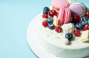 Tender white cake decorated with melted white chocolate, macaroons, meringues, berries and candies on blue background.