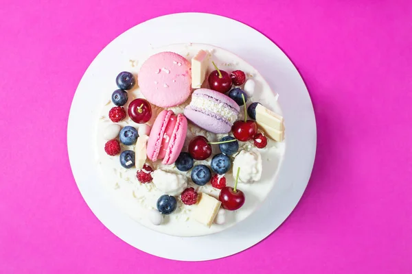 Tender white cake decorated with melted white chocolate, macaroons, meringues, berries and candies on pink background. Top view