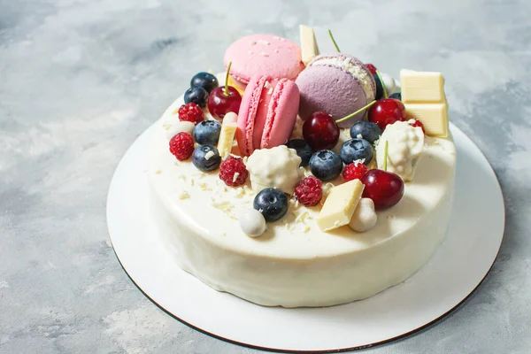 Tender white cake decorated with melted white chocolate, macaroons, meringues, berries and candies on concrete background.