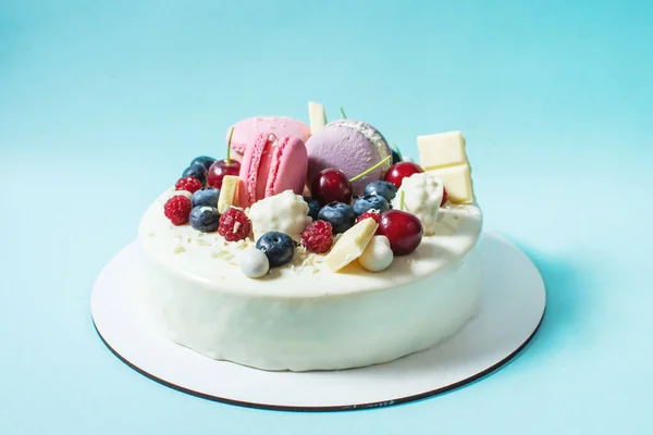 Tender white cake decorated with melted white chocolate, macaroons, meringues, berries and candies on blue background.