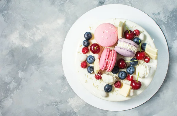 Tender white cake decorated with melted white chocolate, macaroons, meringues, berries and candies on concrete background. Top view