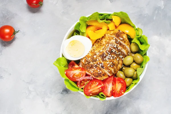 Buddha bowl salad with grilled chicken breast slices, cherry tomatoes, eggs, pepper, olive, lettuce and seeds on concrete background. Healthy food, clean eating, dieting, top view