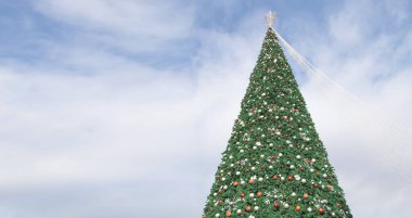 Açık havada mavi bulutlu gökyüzünün altında kırmızı süslemelerle süslenmiş Noel ağacı.