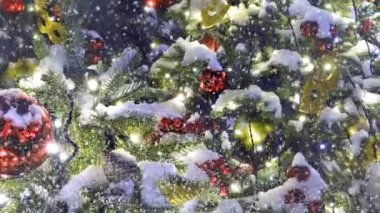 Noel ağacı dışarıda, düşen ve dönen karın altında oyuncaklar ve çelenklerle süslenir. Yeni yıl renkli bir geçmiş. Canlandırılmış 4K video.