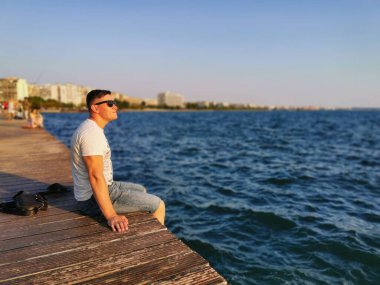 Deniz kenarındaki setin kenarında oturan genç, atletik bir adamın portresi. Gün batımını ve doğayı sever. Seyahat, tatil, Yunanistan