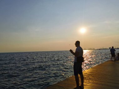 Deniz kenarındaki setin kenarında oturan genç, atletik bir adamın portresi. Gün batımını ve doğayı sever. Seyahat, tatil, Yunanistan