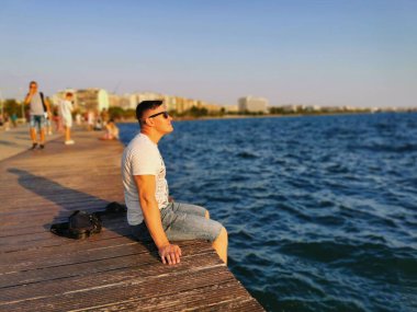 Deniz kenarındaki setin kenarında oturan genç, atletik bir adamın portresi. Gün batımını ve doğayı sever. Seyahat, tatil, Yunanistan