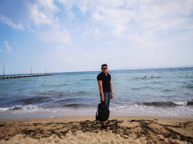 Genç bir adam sahilde sırt çantasıyla duruyor. Uzaktan çalışma, seyahat, eğlence turizmi, Yunanistan plajları, işten dinlenme, uzak çalışma