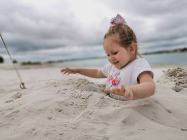 Küçük bir çocuk kumsalda kumsalda oynuyor, kafasına kadar gömülmüş, eşarplı bir kız oyun oynarken gülümsüyor, beyaz kum, yaz, mavi gökyüzü, yatay fotoğraf, aile tatili, çocuklarla oyunlar.