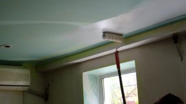 Evin içinde tamirat yapan bir adam tavanı uzun bir sopayla silindirle boyuyor. Tavan beyaz boyayla kaplıydı. Su emülsiyon kaplaması, akrilik yenilemeli eski bir yenileme.