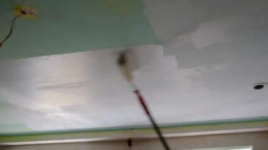 A man makes repairs inside the house, painting the ceiling with a long stick with a roller. Ceiling covered with white paint. Water emulsion coating, renovation of an old renovation with acrylic