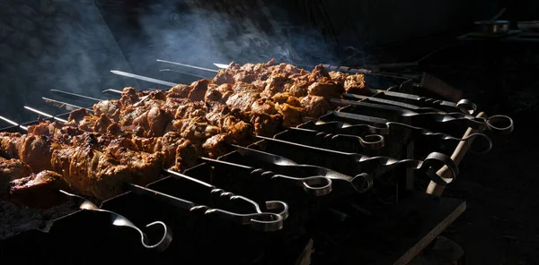 Large pieces of pork meat are fried on coals in the grill. Smoke from coal, dark background.Skewers with strung shish kebab. Fatty delicious food. Food for a large company for a picnic in the forest