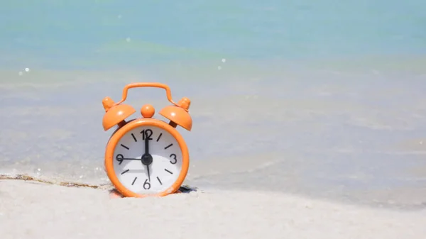 Orange alarm clock on the beach, sand and azure water. Concept: vacation time, tourist season by the water, beach time. copy space, summer banner.