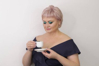 A beautiful elderly woman with bright makeup stands with a cup of hot coffee on a light background. Concept: business woman, coffee break. Press conference, entrepreneur, coffee divination.