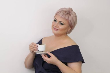 A beautiful elderly woman with bright makeup stands with a cup of hot coffee on a light background. Concept: business woman, coffee break. Press conference, entrepreneur, coffee divination.