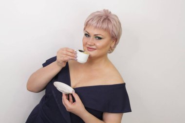 A beautiful elderly woman with bright makeup stands with a cup of hot coffee on a light background. Concept: business woman, coffee break. Press conference, entrepreneur, coffee divination.