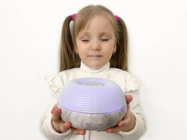 A little funny girl with ponytails inhales essential oils from a diffuser from a humidifier. Device for purification and fresh air. Light background