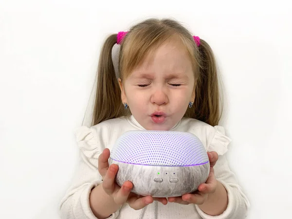 A little funny girl with ponytails inhales essential oils from a diffuser from a humidifier. Device for purification and fresh air. Light background