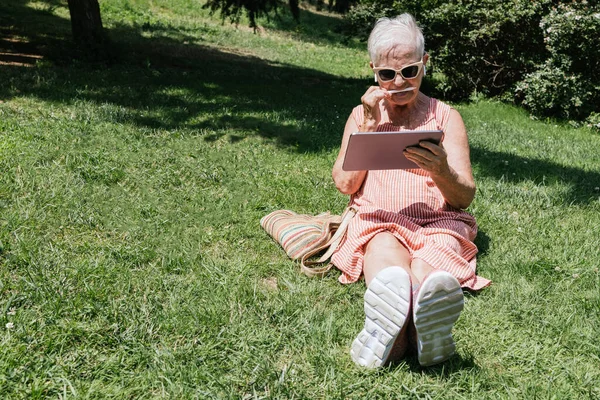 white-haired old lady with a tablet and pencil creating digital art. listening to music on wireless headphones on a summer day.