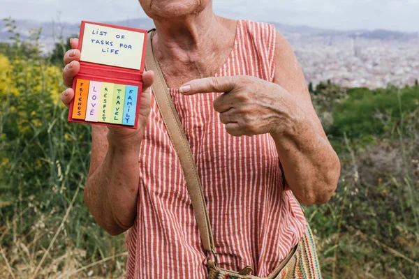 unrecognizable elderly caucasian woman points to a notebook with colored papers with a list of tasks for life.friends,family,travel,respect and love.she has a big city behind her in the background.