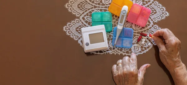 elderly hands holding medication in pill form next to blood pressure monitor and thermometer