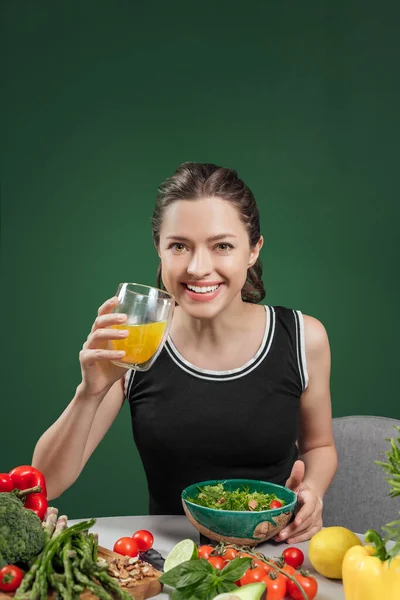Portrait of a young cheerful woman with fresh juice at the table full of healthy raw vegetables and fruits on green background — Stock Photo, Image