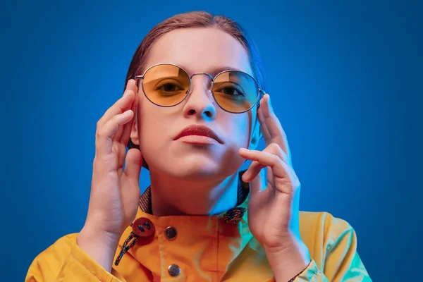 Retrato de niña de moda en impermeable amarillo y gafas de sol redondas en luz de neón sobre fondo azul — Foto de Stock
