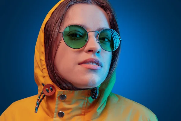 Retrato de niña de moda en impermeable amarillo y gafas de sol redondas en luz de neón sobre fondo azul — Foto de Stock