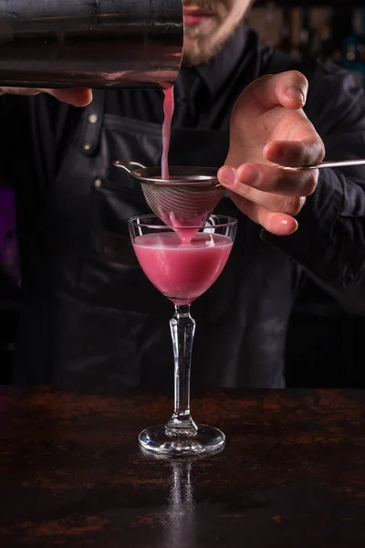 Barman ubrany w czarny fartuch i koszulę zrobić różowy drink koktajlowy na czarnym tle — Zdjęcie stockowe